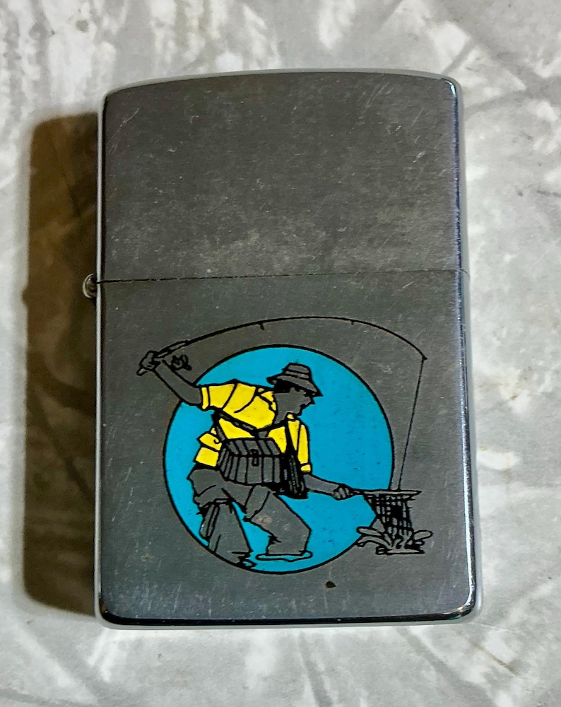 May include: A silver-toned metal lighter with a fishing-themed graphic. The graphic features a fisherman in yellow and gray attire, casting a line against a blue circle backdrop. The lighter has a rectangular shape with a hinged lid.