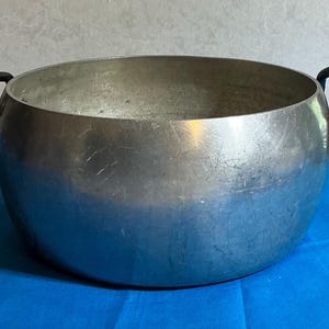 May include: A round, brushed metal bowl with two dark, curved handles. The bowl has a wide opening and a slightly rounded base. The metal has a matte finish and shows some wear. The bowl sits on a blue surface.