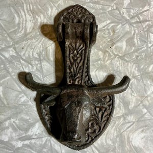 May include: A cast iron wall hook featuring a bull's head design. The hook is dark brown with intricate detailing, including the bull's horns and decorative patterns. The hook is designed to be mounted on a wall.