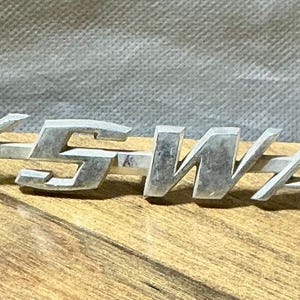 May include: A silver-colored metal emblem with the word "VOLKSWAGEN" in capital letters. The emblem is placed on a wooden surface with a gray fabric background. The letters are blocky and slightly weathered.