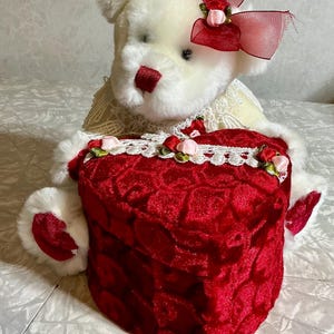 May include: A white teddy bear with a red nose and bow, holding a heart-shaped red velvet box. The box is trimmed with white lace and small pink roses. The bear has a red bow on its head.