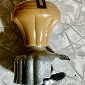 May include: A vintage steering wheel spinner knob featuring a smooth, cream-colored, fluted plastic handle attached to a silver-toned metal clamp mechanism with a textured interior grip designed for mounting onto a steering wheel rim.