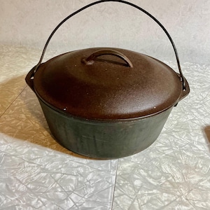 May include: A round cast iron Dutch oven with a dark brown, textured finish. The pot features a matching lid with a central handle and a sturdy, arched metal bail handle attached to the sides for lifting. The surface shows signs of use.