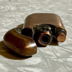 May include: A copper-colored, vintage lighter with a hinged lid and a curved, rounded arm. The lighter has a cylindrical nozzle and a small, textured button. The lighter is open, revealing the interior components.