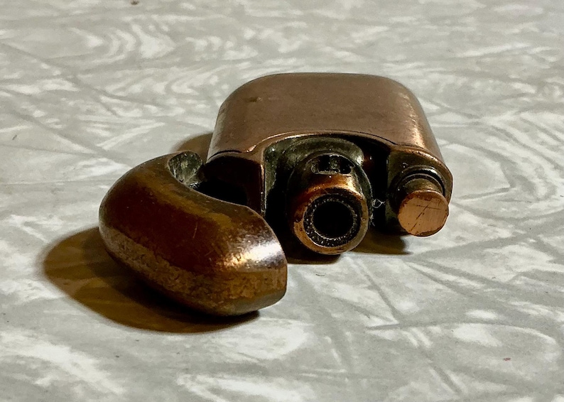May include: A copper-colored, vintage lighter with a hinged lid and a curved, rounded arm. The lighter has a cylindrical nozzle and a small, textured button. The lighter is open, revealing the interior components.
