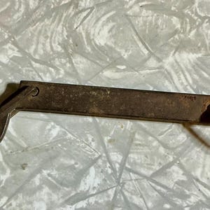 May include: A weathered, dark brown metal latch mechanism featuring a flat, rectangular bar with a hook on one end and a small, oval-shaped metal loop attached to the opposite end. The metal surface shows signs of oxidation and age.