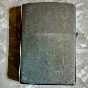 May include: A brushed metal lighter with a rectangular shape. The lighter has a hinged lid and a small metal hinge on the side. The surface of the lighter shows signs of wear with visible scratches.