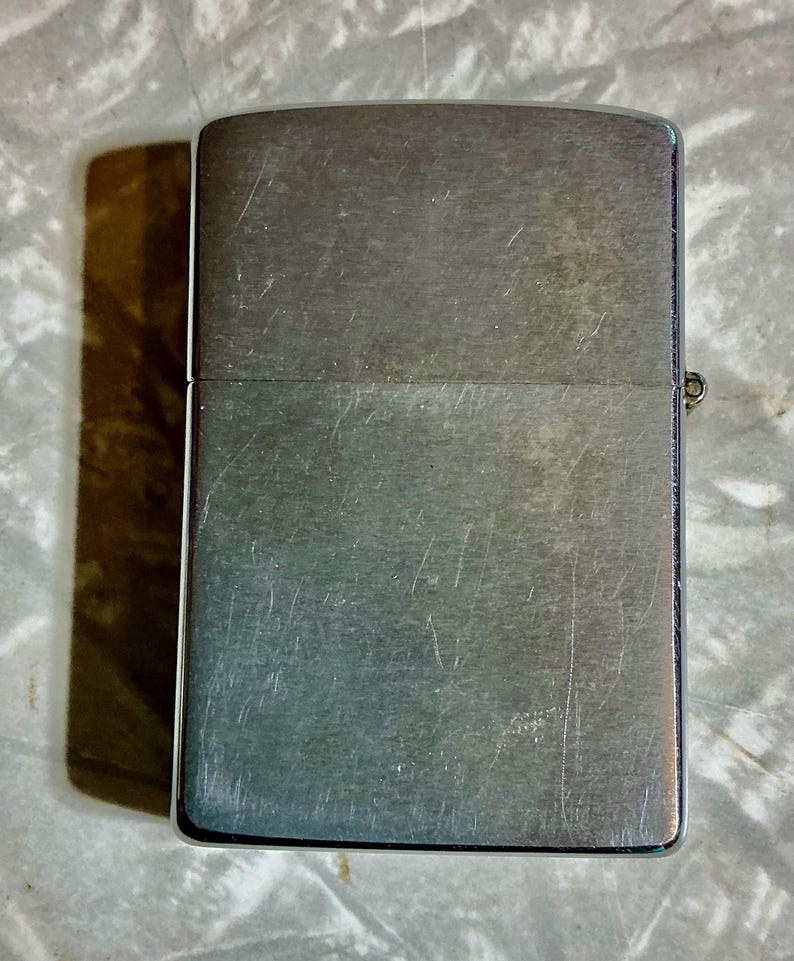 May include: A brushed metal lighter with a rectangular shape. The lighter has a hinged lid and a small metal hinge on the side. The surface of the lighter shows signs of wear with visible scratches.