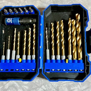 May include: Open blue and black drill bit set with gold drill bits and a blue drill bit holder. The set includes various sizes of drill bits and a drill bit driver. The case is open, revealing the contents.