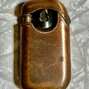 May include: A vintage copper-toned lighter with a rounded rectangular shape. The lighter has a hinged top and a visible flint wheel. The metal shows signs of age and wear, with a patinaed finish. The lighter's design is simple and functional.