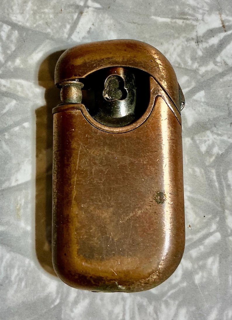 May include: A vintage copper-toned lighter with a rounded rectangular shape. The lighter has a hinged top and a visible flint wheel. The metal shows signs of age and wear, with a patinaed finish. The lighter's design is simple and functional.