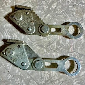 May include: Two silver-colored mini clamps with the text "MINI-CLAMP DEUER MFG. DAYTON, O." are displayed. Each clamp has a pivoting arm and a circular opening at the end. The clamps are on a textured surface.