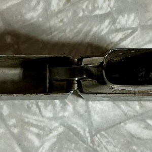 May include: Open, silver-colored lighter with a rectangular shape. The lighter is made of metal and has a hinged lid. The interior of the lighter is visible, showing the fuel compartment and wick. The lighter is resting on a white, textured surface.