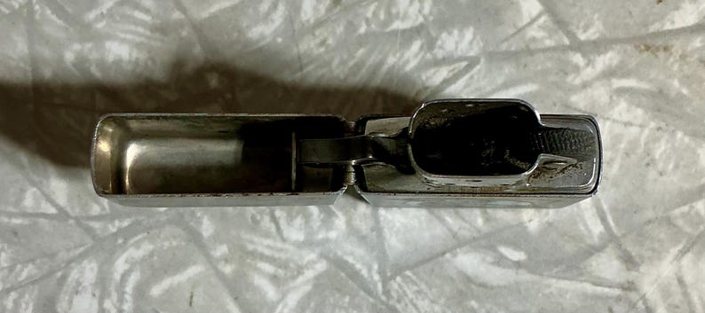 May include: Open, silver-colored lighter with a rectangular shape. The lighter is made of metal and has a hinged lid. The interior of the lighter is visible, showing the fuel compartment and wick. The lighter is resting on a white, textured surface.