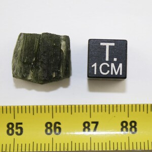 May include: A dark green, rough, and textured rock sample with a 1 cm ruler for scale.