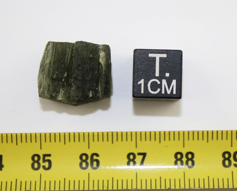 May include: A dark green, rough, and textured rock sample with a 1 cm ruler for scale.