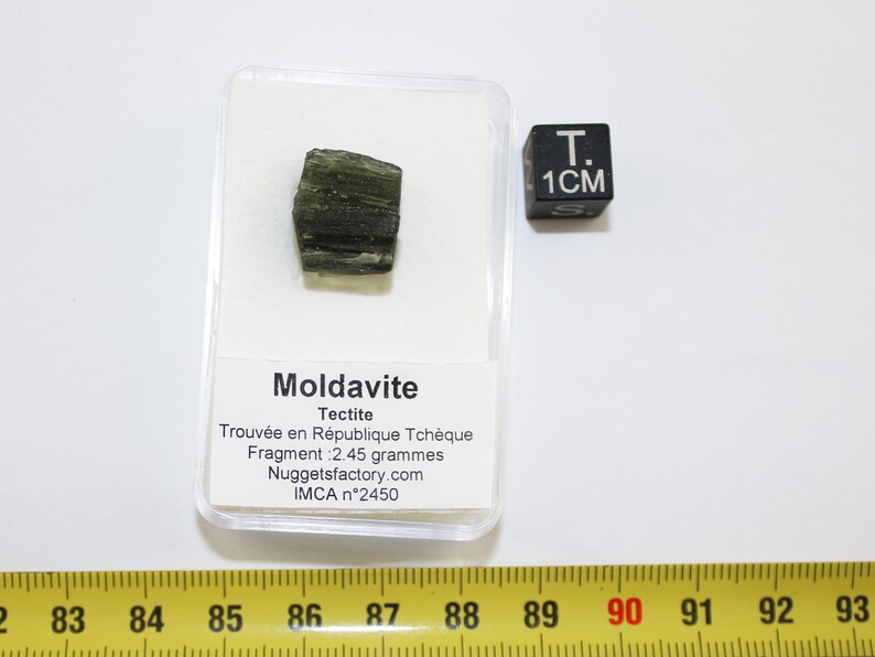 May include: A green moldavite tectite, a type of natural glass, in a clear plastic display case with a white label that reads "Moldavite Tectite Trouv&eacute;e en R&eacute;publique Tch&egrave;que Fragment: 2.45 grammes Nuggetsfactory.com IMCA n&deg;2450". A black cube with the text "T. 1CM" is to the right of the display case.