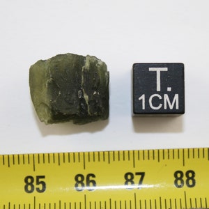 May include: A dark green, rough-textured rock with a slightly translucent quality. The rock is about 1 inch in size, as indicated by the ruler and the 1 cm cube.