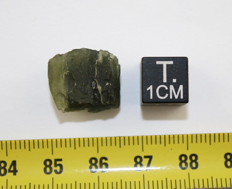 May include: A dark green, rough-textured rock with a slightly translucent quality. The rock is about 1 inch in size, as indicated by the ruler and the 1 cm cube.
