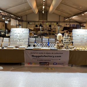 Puede incluir: Un puesto de venta de minerales y f&oacute;siles en una feria de minerales y f&oacute;siles. La mesa tiene un mantel marr&oacute;n y una pancarta que dice "Achat et vente d objets de collection Nuggetsfactory.com &agrave; votre service ebay IMEa catousiki".