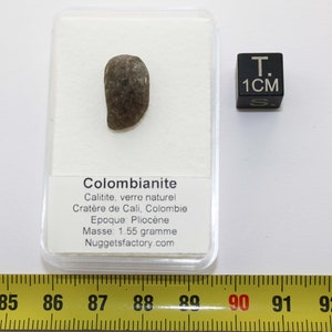 Puede incluir: Un colombianite gris oscuro, un tipo de vidrio natural, en una vitrina de pl&aacute;stico transparente con una etiqueta blanca que dice "Colombianite, Calitite, verre naturel, Crat&egrave;re de Cali, Colombie, Epoque: Plioc&egrave;ne, Masse: 1,55 gramo, Nuggetsfactory.com". Un cubo negro con el texto "T. 1CM S" est&aacute; a la derecha de la vitrina. Una cinta m&eacute;trica amarilla con marcas negras est&aacute; en la parte inferior de la imagen.