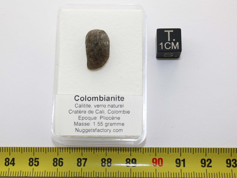 Puede incluir: Un colombianite gris oscuro, un tipo de vidrio natural, en una vitrina de pl&aacute;stico transparente con una etiqueta blanca que dice "Colombianite, Calitite, verre naturel, Crat&egrave;re de Cali, Colombie, Epoque: Plioc&egrave;ne, Masse: 1,55 gramo, Nuggetsfactory.com". Un cubo negro con el texto "T. 1CM S" est&aacute; a la derecha de la vitrina. Una cinta m&eacute;trica amarilla con marcas negras est&aacute; en la parte inferior de la imagen.