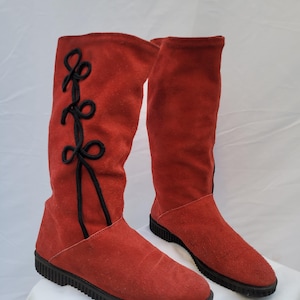May include: Red suede knee-high boots with black trim and decorative laces. The boots have a flat sole and a textured black rubber sole. The laces are a black decorative element on the side of the boots.