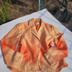Puede incluir: Una blazer color melocotón con acentos degradados en naranja. La blazer presenta un cierre de un solo botón, un dobladillo con volantes y un pequeño bolsillo. La etiqueta "Tuzzi" es visible en el cuello. Las mangas son largas y con puños.