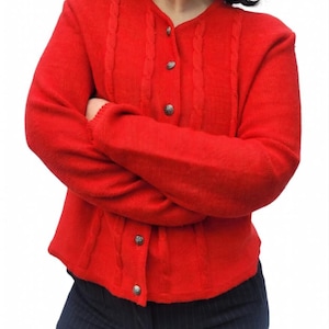 Puede incluir: Un cárdigan rojo brillante con cuello redondo y botones plateados. El suéter presenta un diseño de punto trenzado en la parte delantera y en las mangas. La persona lleva pantalones de rayas finas azul oscuro.