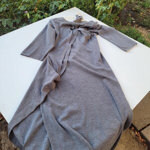 May include: A gray, long-sleeved dress with a bow detail at the neckline. The dress is made of a soft fabric and has a flowing design. The dress is displayed on a white surface.