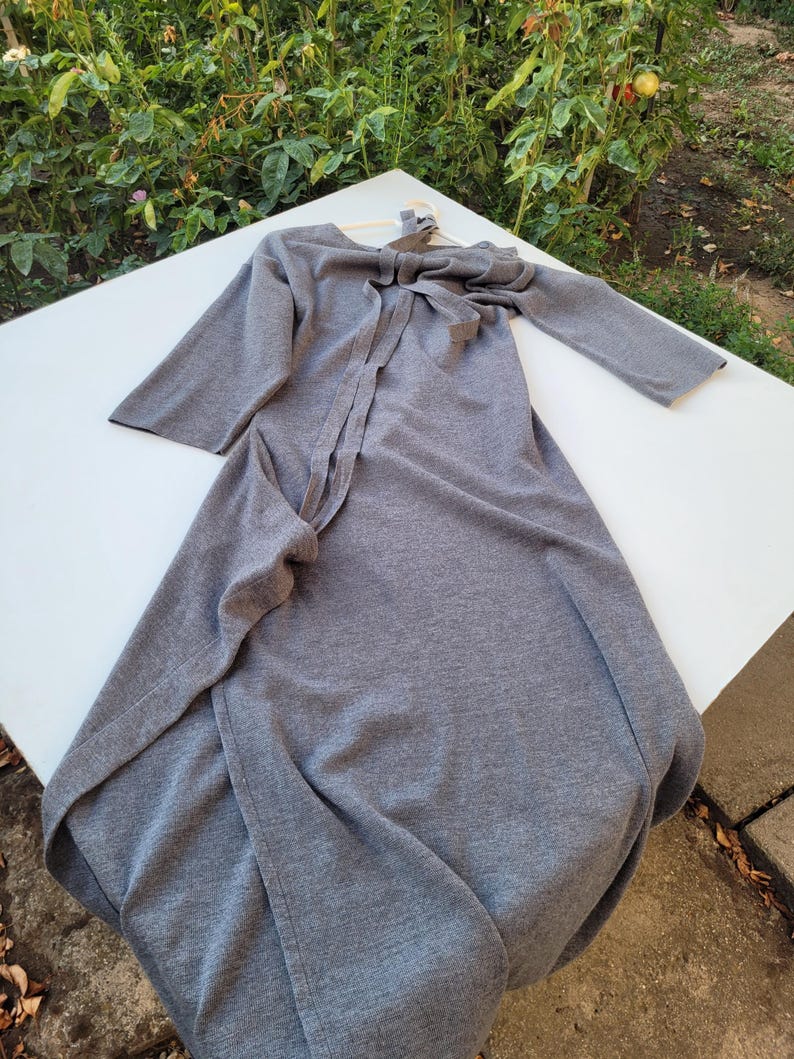 May include: A gray, long-sleeved dress with a bow detail at the neckline. The dress is made of a soft fabric and has a flowing design. The dress is displayed on a white surface.