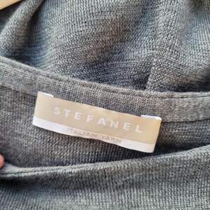 May include: Close-up of a gray knit sweater with the brand label "STEFANEL ITALIAN YARN" visible. The sweater is hanging on a wooden hanger. The fabric has a fine, textured weave.