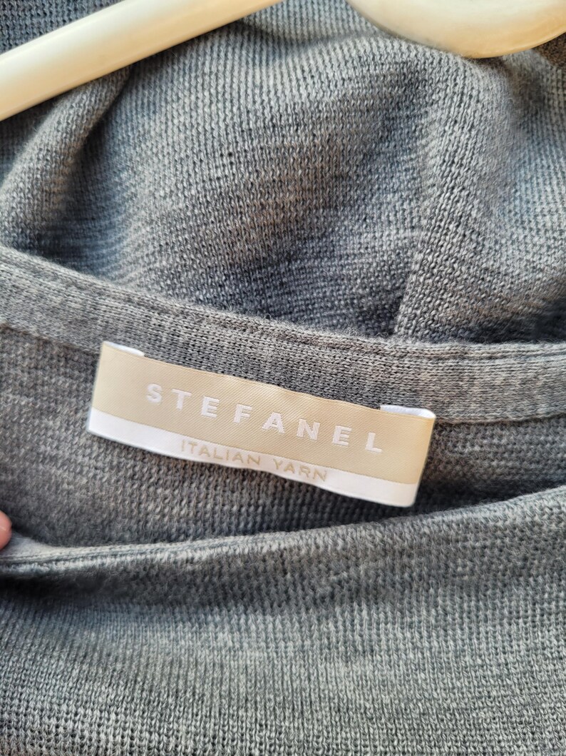 May include: Close-up of a gray knit sweater with the brand label "STEFANEL ITALIAN YARN" visible. The sweater is hanging on a wooden hanger. The fabric has a fine, textured weave.