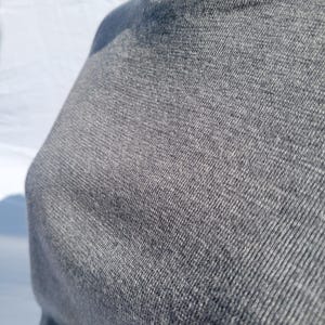 May include: Close-up of a gray, crew neck sweater. The fabric has a textured appearance, with visible woven threads. The sweater is a solid color, and the neckline is visible. The garment appears to be made of a soft, comfortable material.