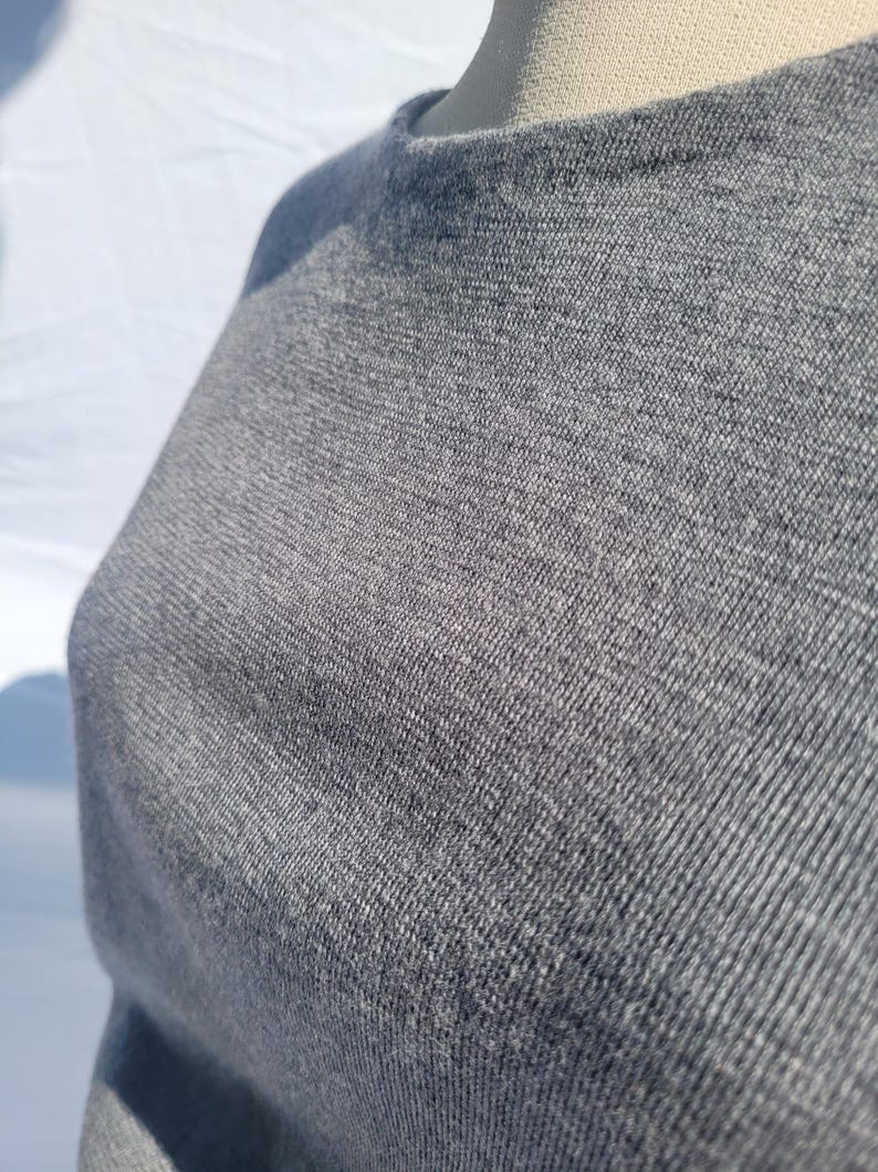 May include: Close-up of a gray, crew neck sweater. The fabric has a textured appearance, with visible woven threads. The sweater is a solid color, and the neckline is visible. The garment appears to be made of a soft, comfortable material.