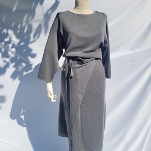 May include: A gray dress displayed on a mannequin. The dress features long sleeves, a wrap-style skirt, and a tie at the waist. The dress is made of a soft, knit fabric. The mannequin is on a white surface.