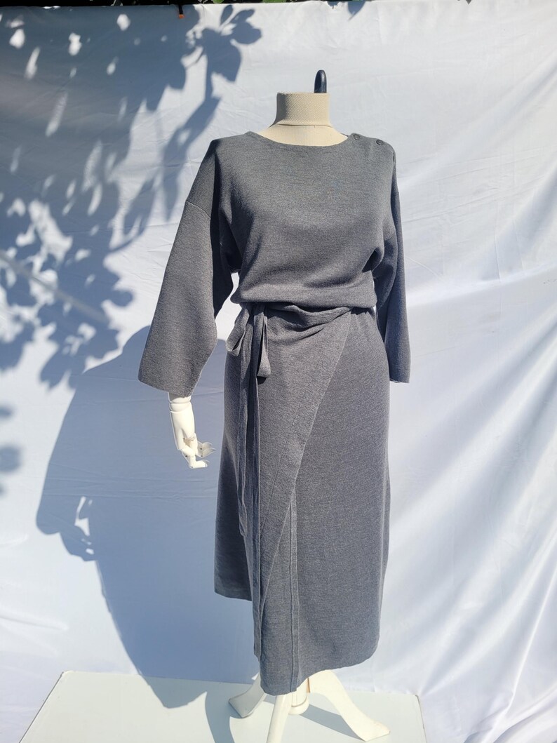 May include: A gray dress displayed on a mannequin. The dress features long sleeves, a wrap-style skirt, and a tie at the waist. The dress is made of a soft, knit fabric. The mannequin is on a white surface.