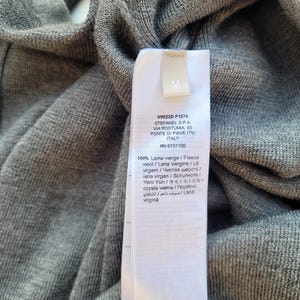 May include: Close-up of a gray knit garment with a white tag. The tag has the letter "M" and text including "VW023D F1274" and "STEFANEL S.P.A." The garment is made of 100% virgin wool.