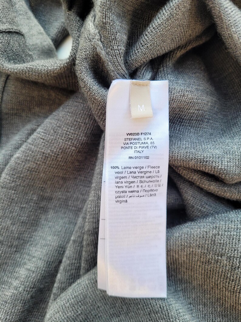 May include: Close-up of a gray knit garment with a white tag. The tag has the letter "M" and text including "VW023D F1274" and "STEFANEL S.P.A." The garment is made of 100% virgin wool.