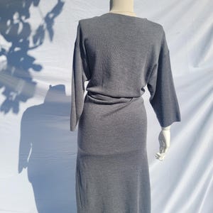 May include: A gray dress with long sleeves and a midi-length skirt is displayed on a mannequin. The dress has a simple, elegant design with a cinched waist. The mannequin is set against a white backdrop, with shadows from foliage adding visual interest.