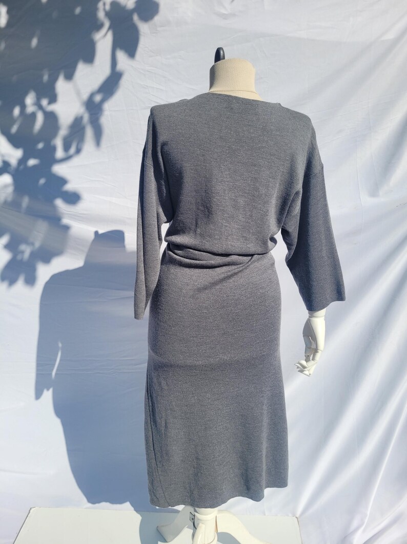 May include: A gray dress with long sleeves and a midi-length skirt is displayed on a mannequin. The dress has a simple, elegant design with a cinched waist. The mannequin is set against a white backdrop, with shadows from foliage adding visual interest.