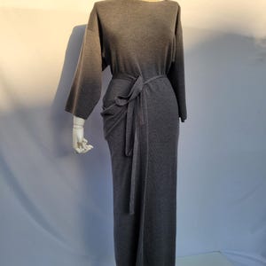 May include: A gray, long-sleeved dress with a boat neck and a tie belt. The dress is displayed on a mannequin. The fabric appears to be a soft knit material. The dress has a long, straight skirt.