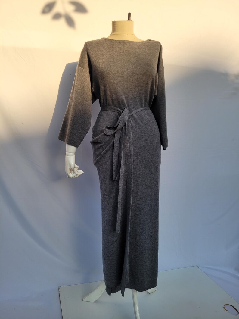 May include: A gray, long-sleeved dress with a boat neck and a tie belt. The dress is displayed on a mannequin. The fabric appears to be a soft knit material. The dress has a long, straight skirt.