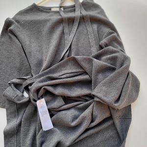 May include: A gray, long-sleeved dress with a tie detail at the neckline and waist, hanging on a white hanger. The dress is made of a soft, knit fabric. A white tag is attached to the dress.