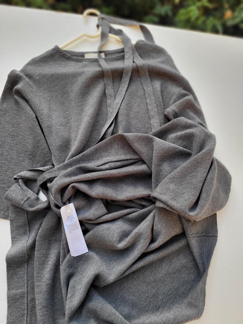 May include: A gray, long-sleeved dress with a tie detail at the neckline and waist, hanging on a white hanger. The dress is made of a soft, knit fabric. A white tag is attached to the dress.