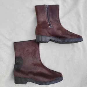 Pode incluir: Um par de botins castanhos com uma aparência texturizada, semelhante a pele. Uma bota tem fecho de correr. As botas têm sola escura e salto baixo, adequadas para o uso diário.