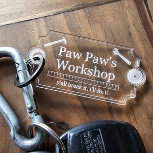 Könnte beinhalten: Ein transparenter Schlüsselanhänger aus Acryl mit dem Text "Paw Paw's Workshop" und dem Spruch "y'all break it, I'll fix it". Der Schlüsselanhänger zeigt ein Lineal, einen Hammer, einen Schraubenschlüssel und eine Säge.
