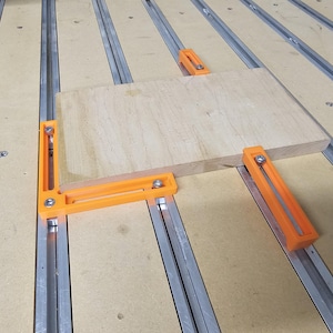 Peut inclure: Une pièce de bois est fixée à un établi à l'aide de quatre serre-joints d'angle orange. Les serre-joints sont fixés à un système de rails sur l'établi.
