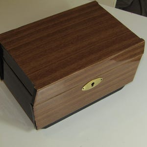 Peut inclure: Une boîte de rangement rectangulaire avec une finition en grain de bois brun foncé sur le couvercle et le panneau avant. La boîte comprend une plaque de serrure ovale en métal de couleur laiton sur le devant. La base et les côtés sont noirs.
