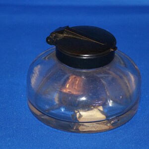 Peut inclure: Un encrier en verre transparent avec une base large et arrondie et un couvercle noir à charnière. Le récipient présente un corps lisse et translucide permettant de voir l'intérieur, surmonté d'un capuchon circulaire sombre avec un petit levier.